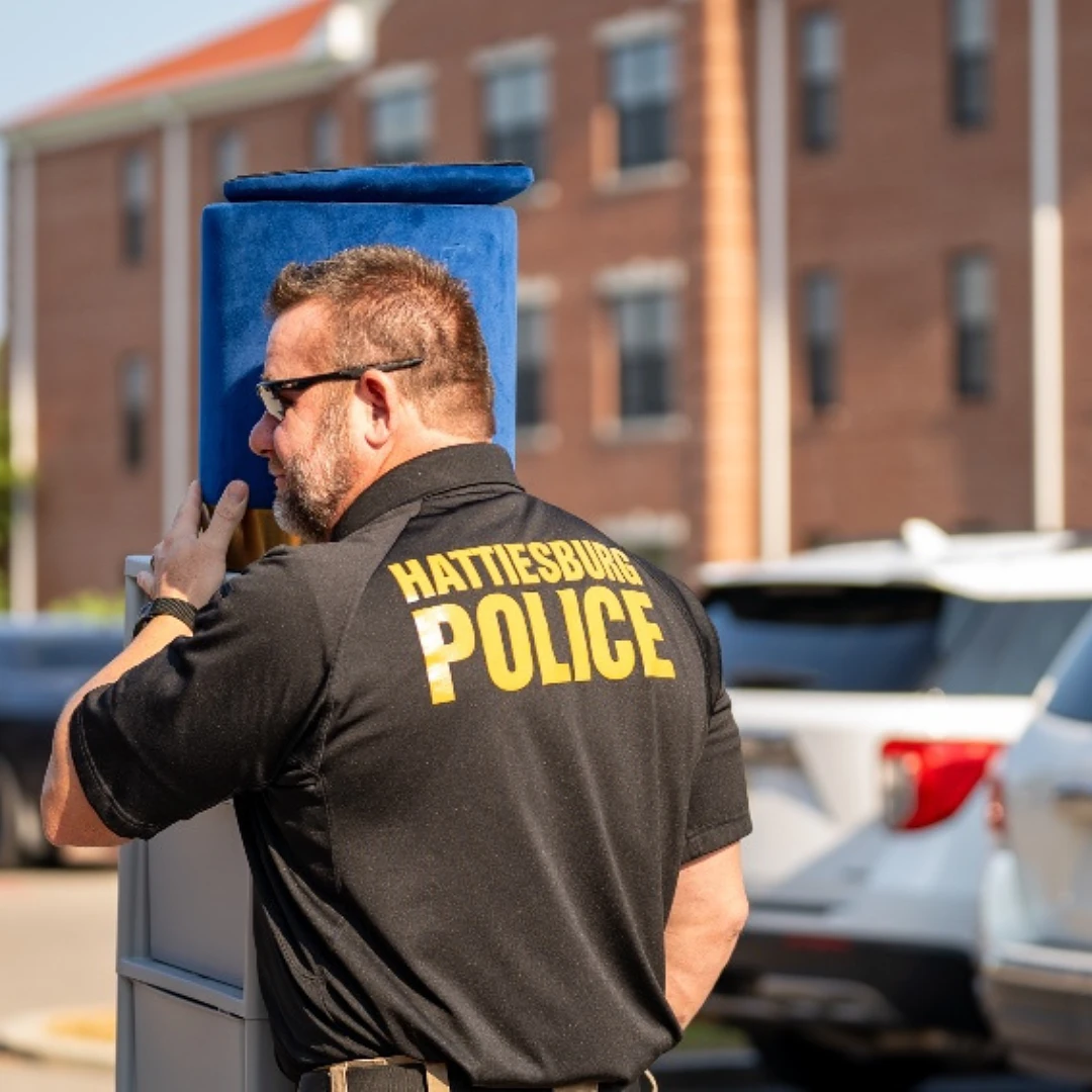 A man in a "Hattiesburg Police" shirt is seen carrying a blue object outdoors near parked cars and buildings.